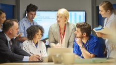 Several people standing and sitting around a table with a screen in the background that has a graph on it