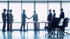 Group of three people on left, two people shaking hands in middle, three people standing behind office chairs on right