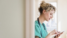 Healthcare provider standing in a hallway wearing green scrubs and a stethoscope around their neck while looking at a tablet