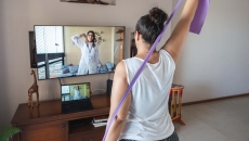 A woman watching and copying exercises with a resistance band in her living room, guided by a physical therapist online