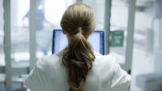 Woman doctor with long red hair using laptop in hospital, photographed from behind