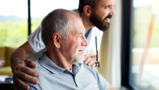 Person sitting in a chair looking out a window with a healthcare provider next to them with their hands on the person's shoulders