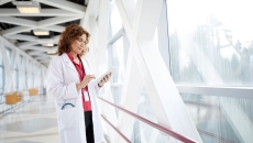 Healthcare provider holding a tablet while wearing a lab coat and standing in a hallway by a large window