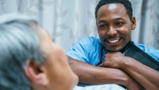 Healthcare provider sitting beside a patient's bed with the patient in it while the provider is holding a tablet