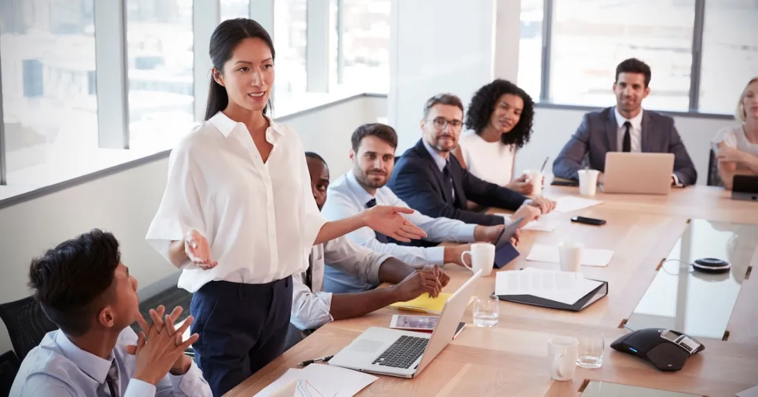 Person standing up at a table in a meeting room with other people sitting down and looking at them