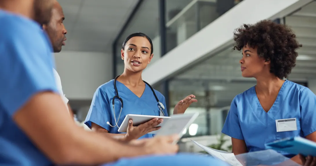 Four healthcare professionals wearing blue scrubs and sitting in a circle with one holding a tablet
