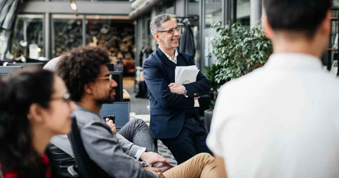 A person standing up, holding paperwork watching something while two other people are sitting and one other person has their back turned