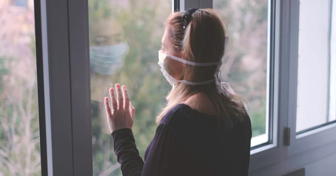 Person with a mask on standing by a window looking out