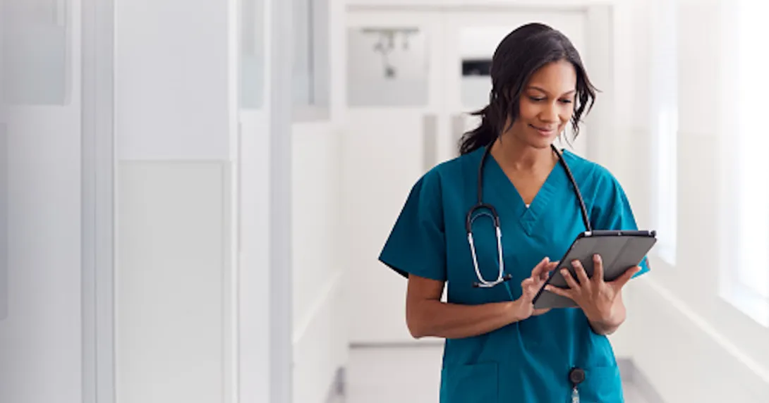 Nurse walking down a white hallway while wearing scrubs and looking at a tablet