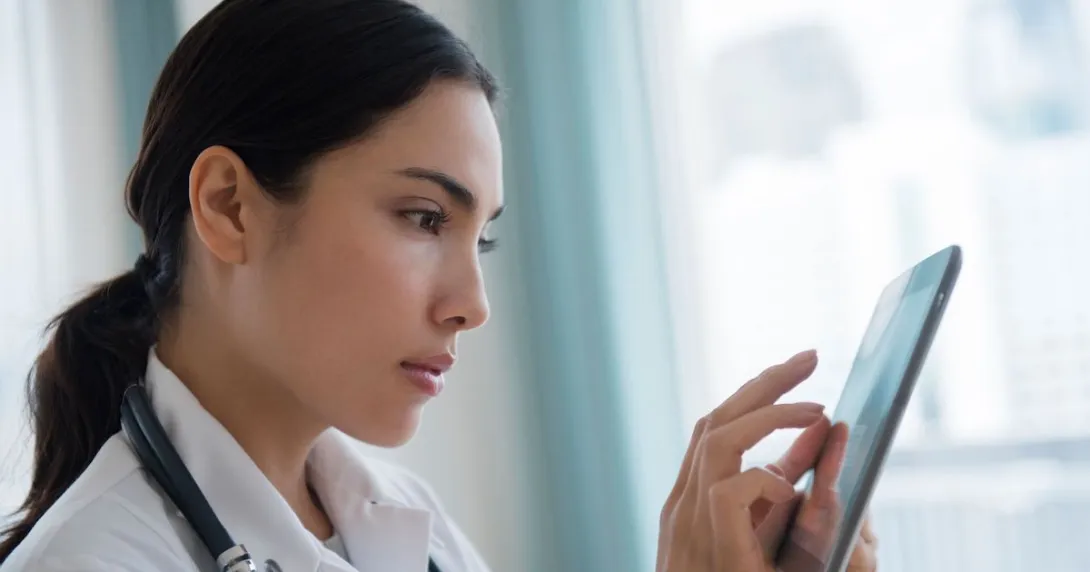 Healthcare provider wearing a lab coat with a stethoscope around their neck and looking at a tablet
