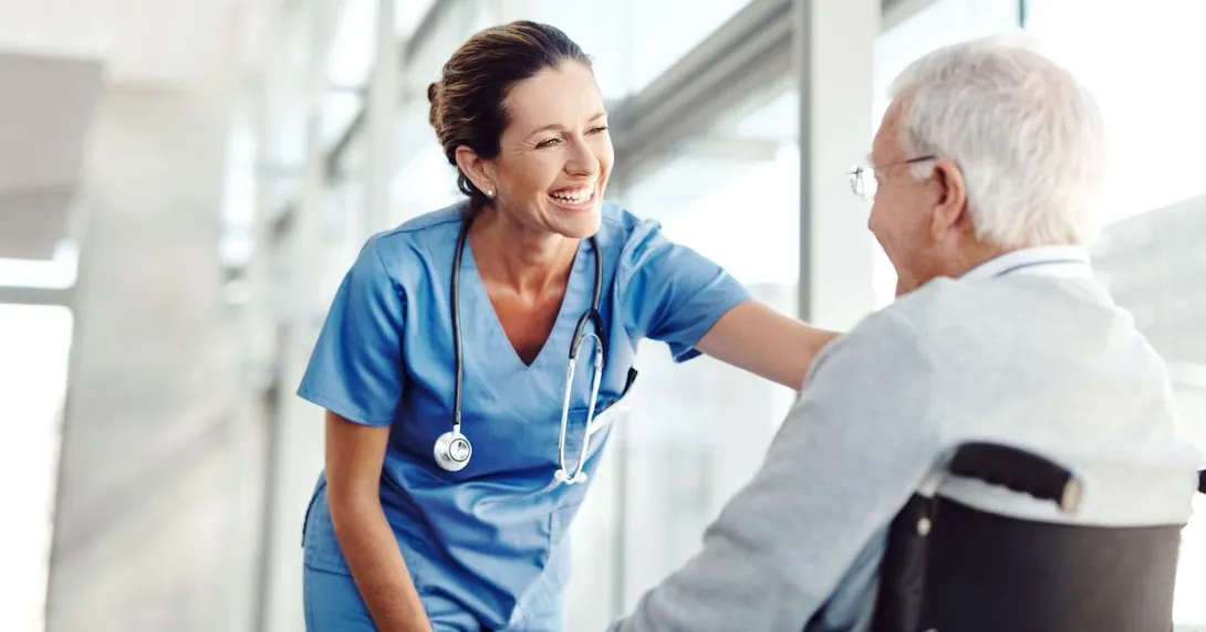 A nurse smiling and standing in front of a window leaning over a patient in a wheelchair