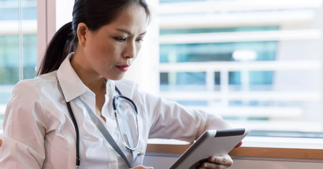 Person sitting by a window wearing a stethoscope and looking at a tablet