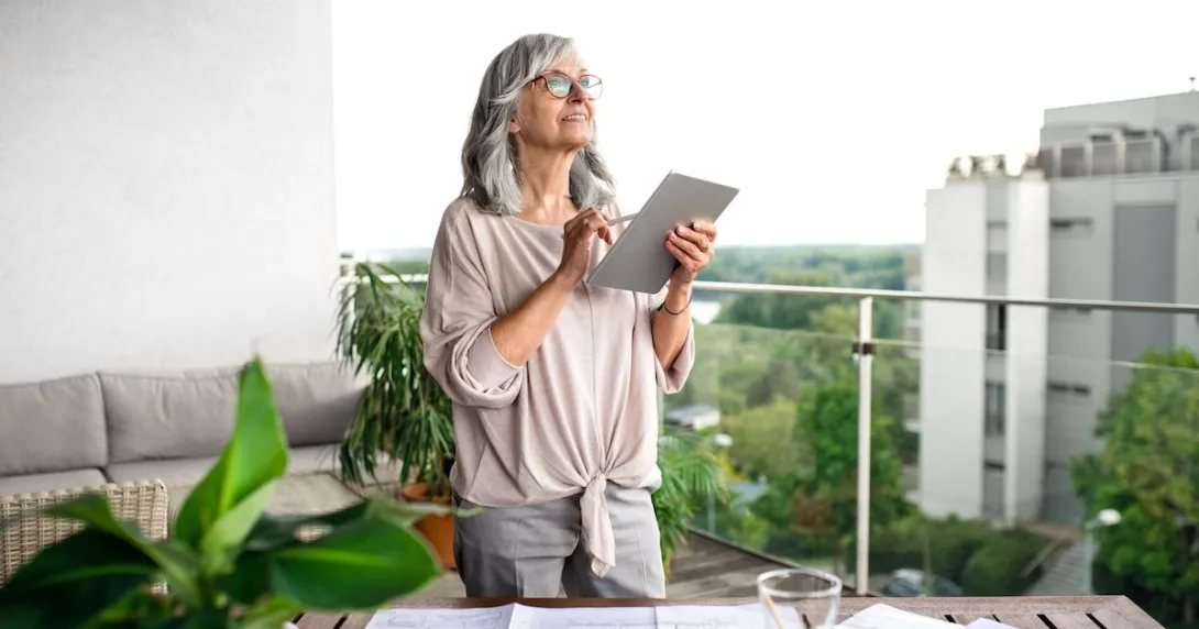 Person standing on a balcony holding a tablet while looking up at the sky with a city behind them