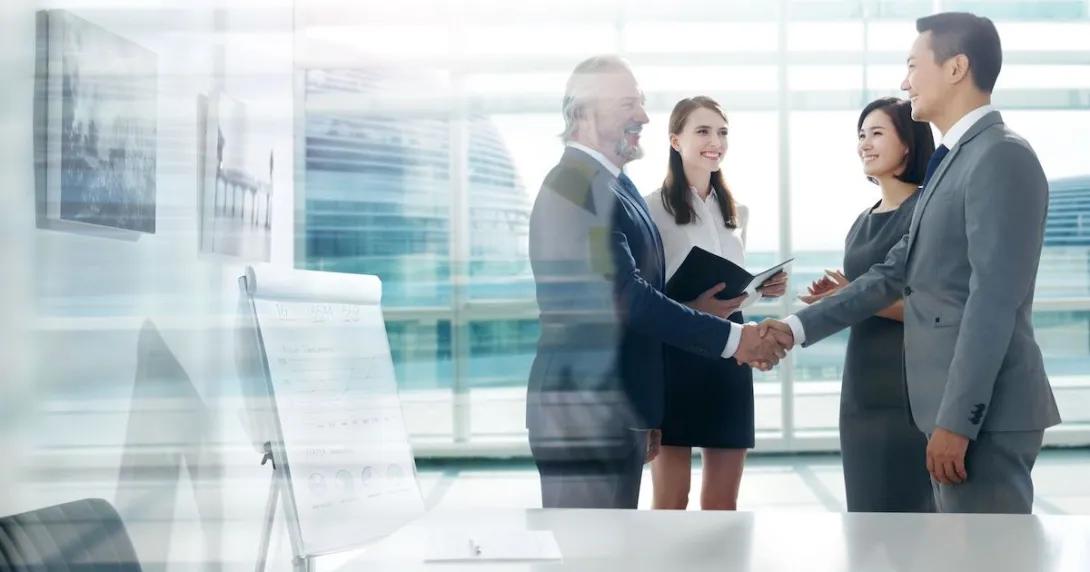 Two people shaking hands with two other people watching, all standing in front of a big window