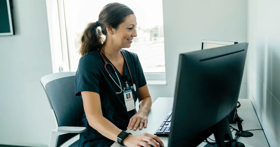 A healthcare provider with scrubs on and a stethoscope around their neck sitting at a desk while looking at a computer