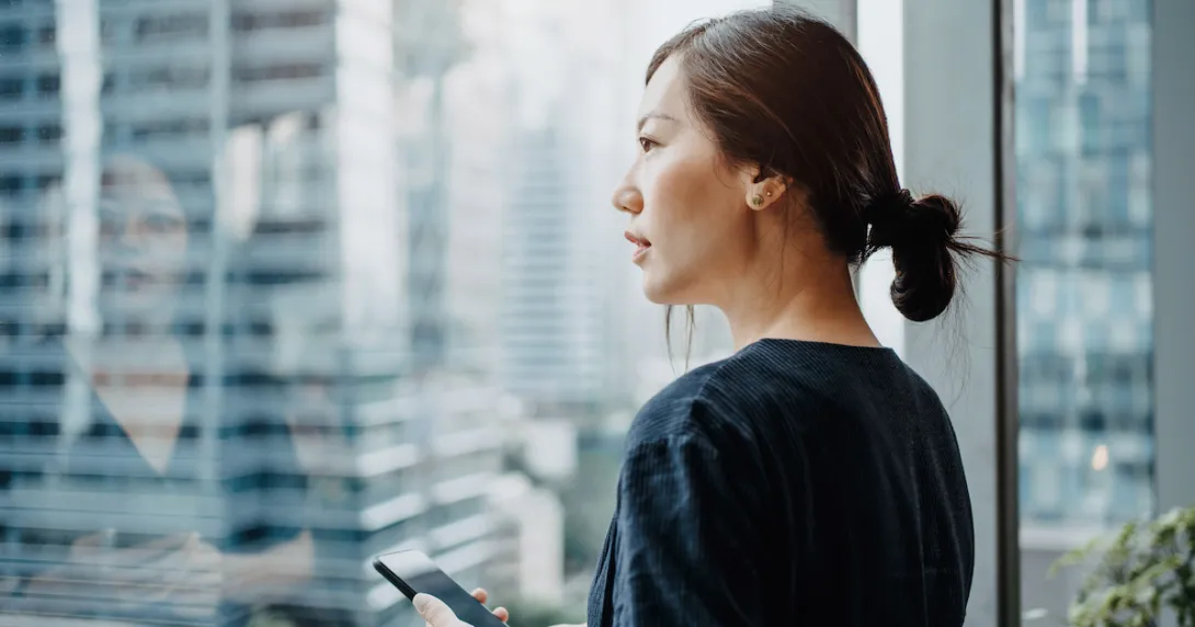 A woman looking out a window holding a smartphone