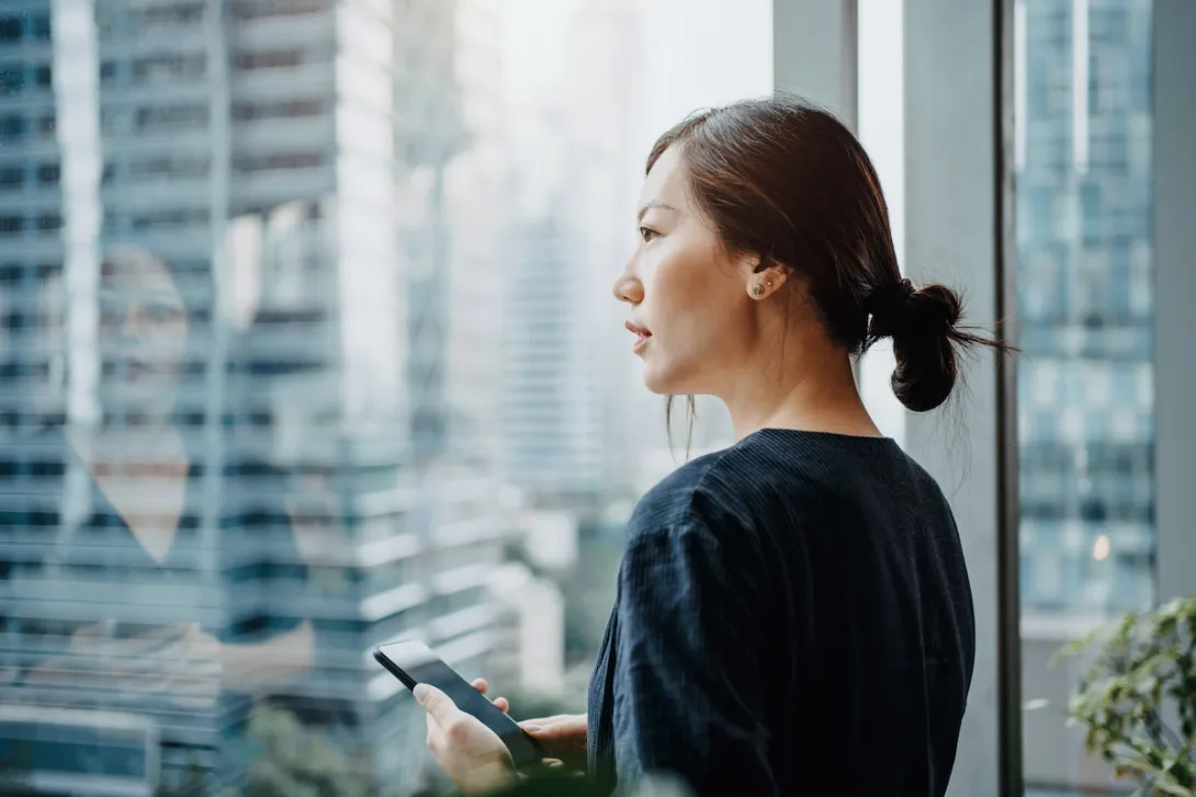 A young woman using a smartphone and looking out a window.
